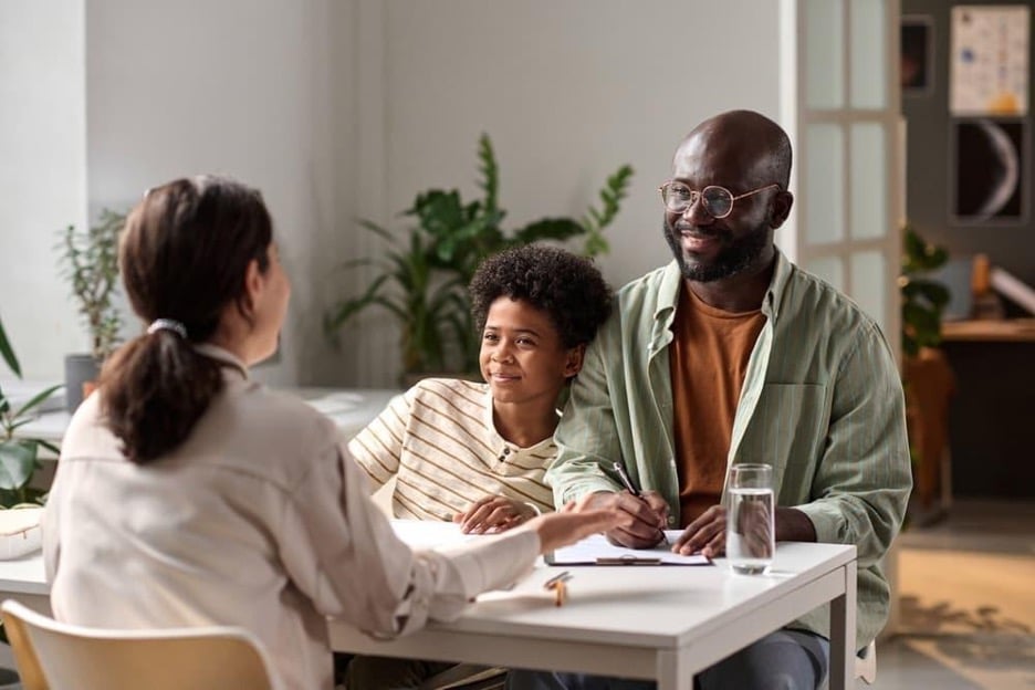 A smiling African American man and his son sit at a desk across from a female social worker