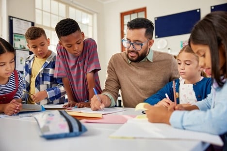 Elementary school students gather around their teacher as he guides them through a writing assignment.